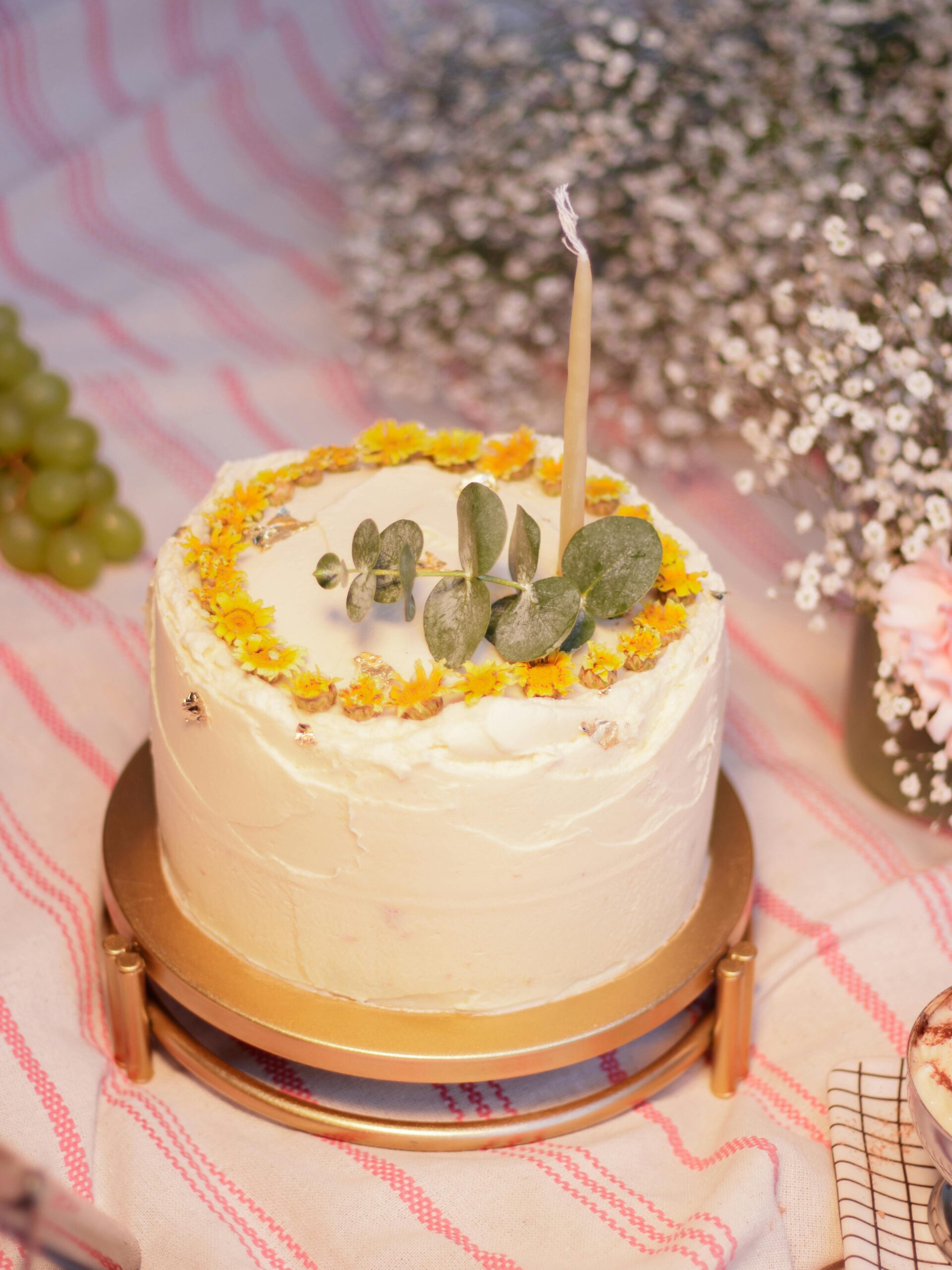 A beautifully decorated floral cake with a single candle on a gold stand, surrounded by delicate flowers.